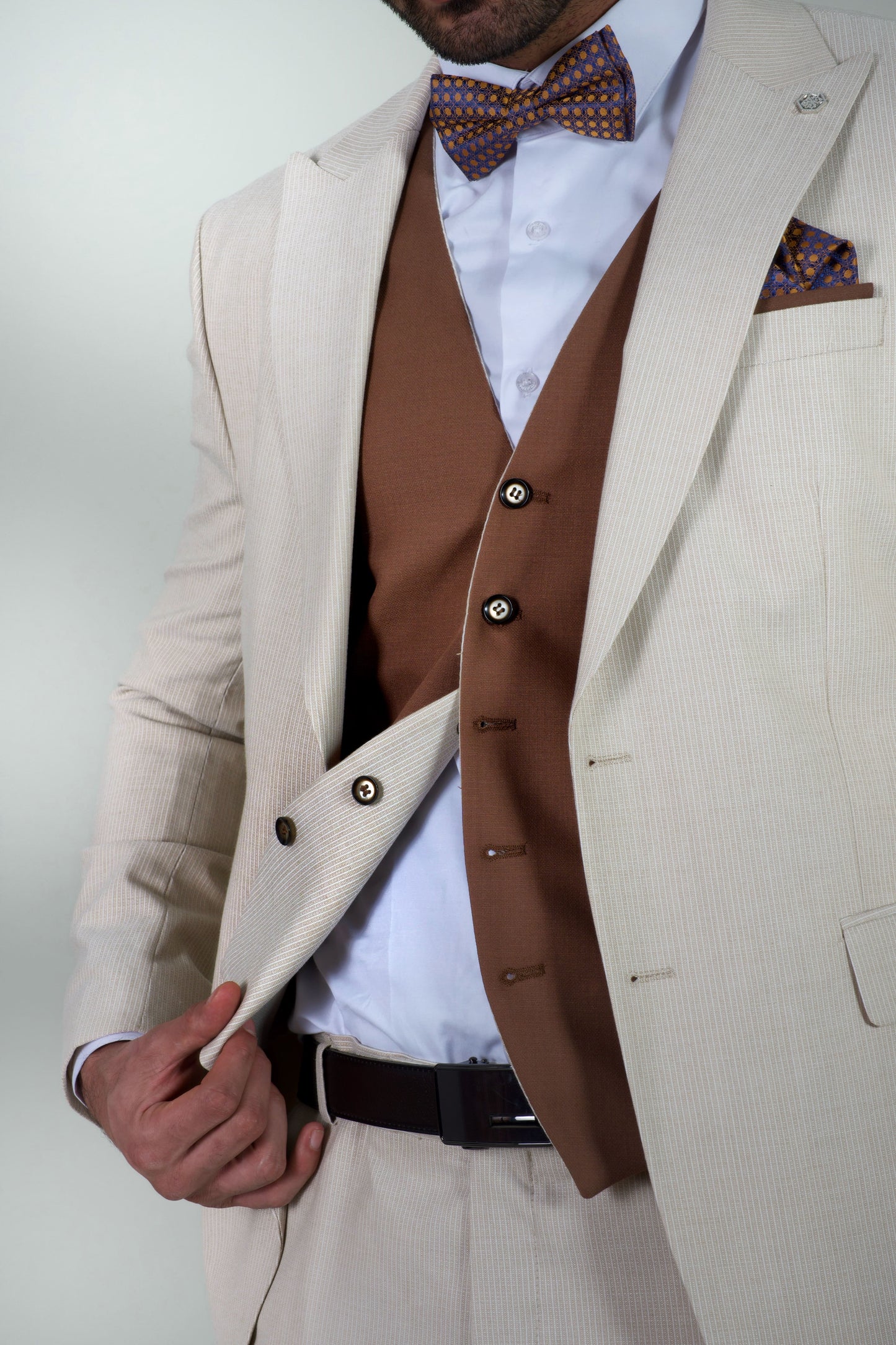 Елегантен мъжки костюм от три части в светлобежов цвят с вталена slim fit кройка, кафява жилетка и папийонка, подходящ за сватба или бал.