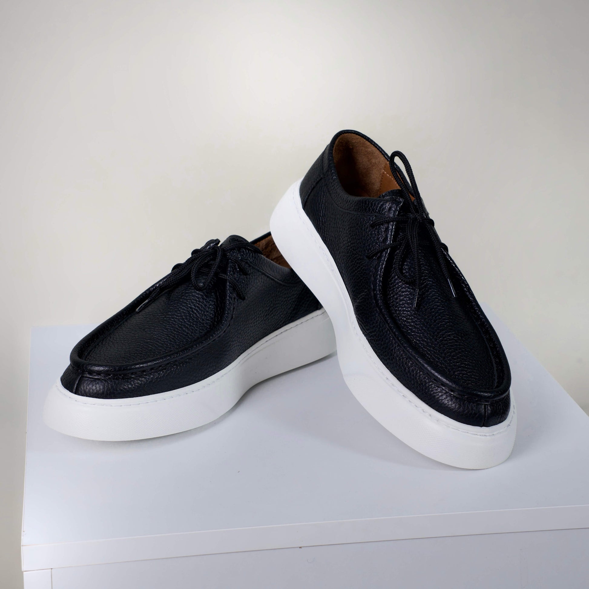 Модерни мъжки обувки от черна текстурирана естествена кожа с висок борд, бели подметки и кожен хастар, заснети върху бял пиедестал.