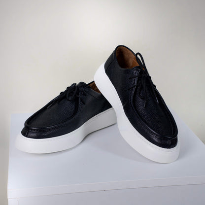 Модерни мъжки обувки от черна текстурирана естествена кожа с висок борд, бели подметки и кожен хастар, заснети върху бял пиедестал.